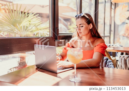Woman works on laptop in a sunny cafe, glass of juice nearby. The setting is modern, with natural light illuminating the space. The image captures a moment of focused work. Woman works on laptop in a sunny cafe, glass of juice nearby. The setting is modern, with natural light illuminating the space. The image captures a moment of focused work. 113453013