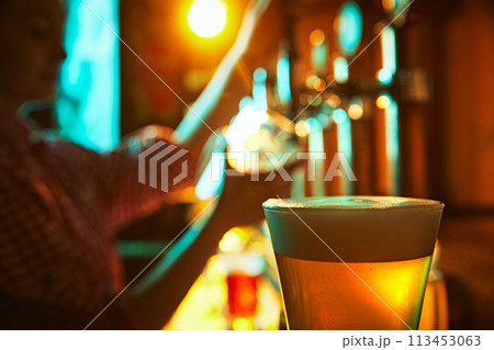 Focus on glass with foamy lager beer and blurred image of bartender pouring another glass of beer in pub on background. 113453063