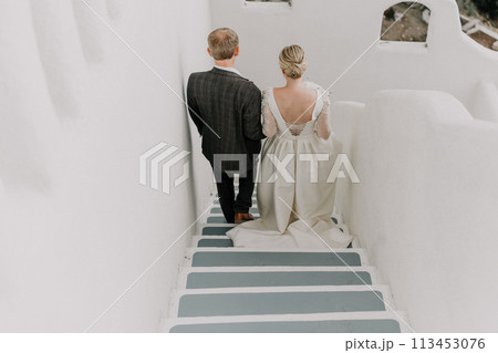 A bride and groom are walking down a white staircase. The bride is wearing a white dress and the groom is wearing a suit. A bride and groom are walking down a white staircase. The bride is wearing a white dress and the groom is wearing a suit. 113453076