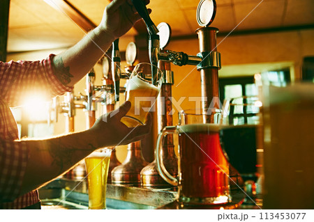 Side view of young bartender pouring beer while standing at the bar counter. Warm pub atmosphere Side view of young bartender pouring beer while standing at the bar counter. Warm pub atmosphere 113453077