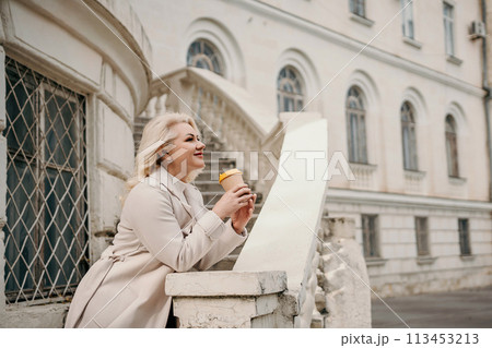 A woman in a white coat is standing on a white railing with a cup of coffee in her hand. She is smiling and looking out at the street. 113453213