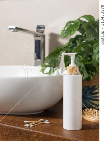 White one pump bottle near basin and green monstera on wooden countertop in bath, mockup 113453579