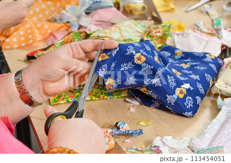 Womans Hands Cutting Blue Fabric with Silver Scissors on Table with Fabric Scraps in Sewing Studio Womans Hands Cutting Blue Fabric with Silver Scissors on Table with Fabric Scraps in Sewing Studio 113454051