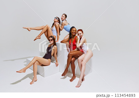 Beautiful woman of different races in stylish swimsuits lounge gracefully on white blocks against white studio background. 113454893
