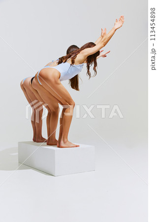 Back view of women in sleek swimsuits stands in diving posture on white platform against white studio background. Back view of women in sleek swimsuits stands in diving posture on white platform against white studio background. 113454898
