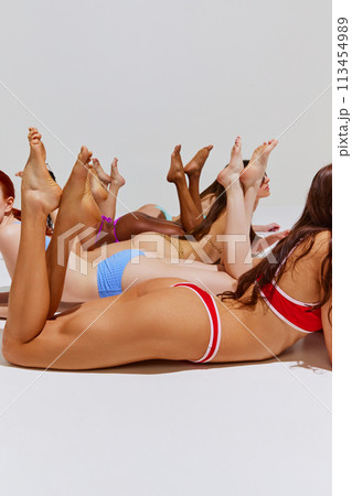 Pretty models in colorful swimwear, slim women of different races posing on floor in bikini against white studio background. 113454989