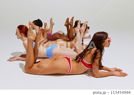 Photo of models in swimwear, beautiful, fit women of different races posing on floor in bikini against white studio background. Photo of models in swimwear, beautiful, fit women of different races posing on floor in bikini against white studio background. 113454994