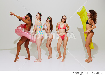 Women wearing colorful bikinis, with sunglasses and pool floats posing with playful expression against white studio background. 113455037