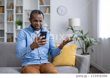 Perplexed African American man sitting in a living room, holding a smartphone and gesturing in confusion over a deceptive message. 113458300