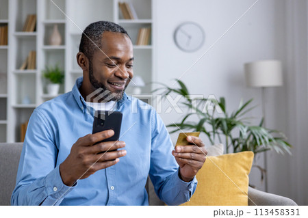 Casual man using smartphone and credit card for online shopping while sitting comfortably at home with indoor plants and bright interior. 113458331