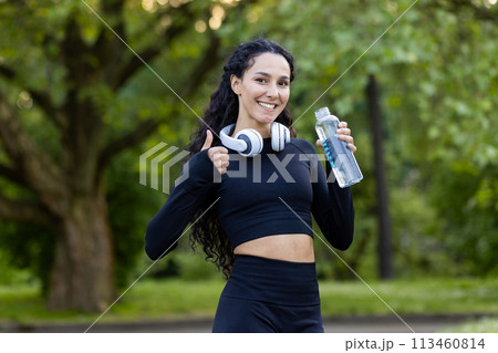 A cheerful Hispanic woman enjoys a fitness break at the park, holding a water bottle and wearing headphones, giving a thumbs up with trees in the background. 113460814