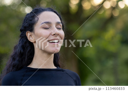 A hispanic woman closes her eyes with a content smile, basking in serenity amidst a natural green backdrop. A hispanic woman closes her eyes with a content smile, basking in serenity amidst a natural green backdrop. 113460833