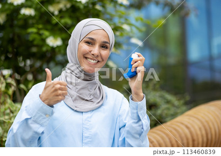A Muslim woman in a hijab holds an asthma inhaler in a park, giving a thumbs up for health and wellness. 113460839