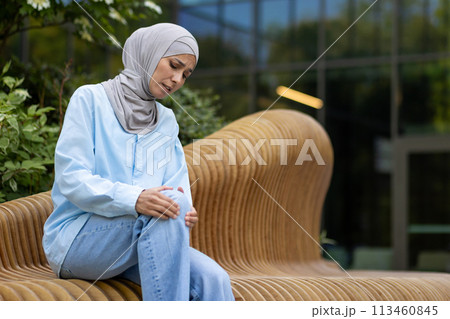 Muslim female sitting outside and raising leg bent in knee with suffering expression on background of modern building. Sick woman facing trauma consequences and rubbing injured place with both hands. Muslim female sitting outside and raising leg bent in knee with suffering expression on background of modern building. Sick woman facing trauma consequences and rubbing injured place with both hands. 113460845
