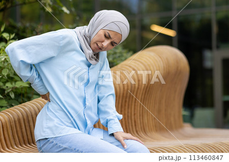 Diverse female person suffering from pain in back or reins due to incorrect posture while spending day outdoors. Unhappy woman feeling sudden acute ache in spinal muscles and leaning forward. 113460847