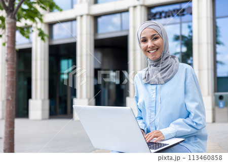 Smiling professional woman wearing a hijab works on her laptop, seated outside an office building with a modern facade. 113460858