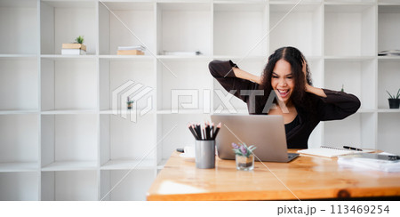 Excited businesswoman with arms raised in a victory gesture, celebrating a successful moment while working on her laptop in the office. 113469254