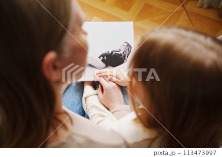 mother and daughter flips through a book with photos of dad and pregnant mom. 113473997