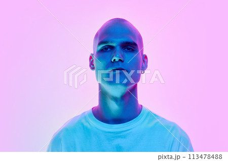 Portrait of serious young man with bald head, shaved face and ear piercing posing on gradient blue pink background in neon light 113478488