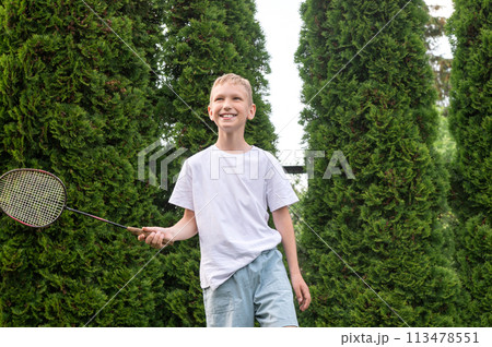 A happy boy holds a badminton racket in his hands, playing sports outside in summer A happy boy holds a badminton racket in his hands, playing sports outside in summer 113478551