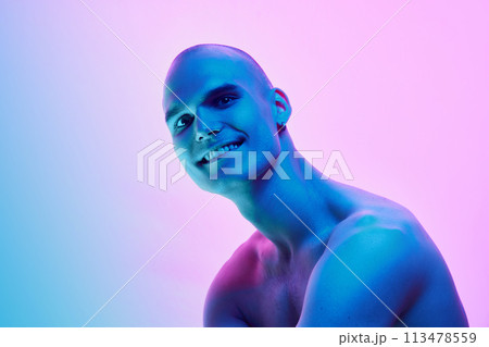 Handsome young man with bald head, shaved face posing shirtless on gradient blue pink background in neon light 113478559