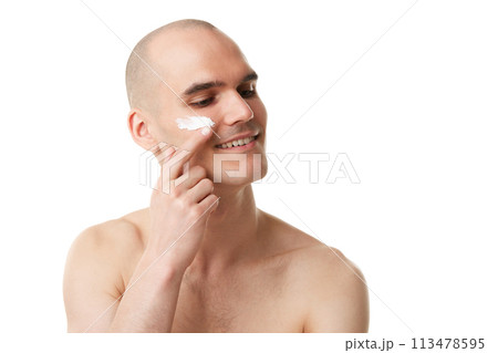 Handsome young bald man taking care after skin, applying face moisturizing cream isolated on white studio background 113478595