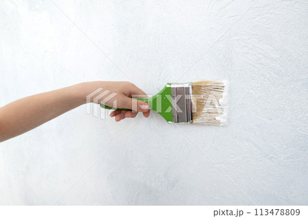 The child's hand holds a paint brush with white paint. Paint the wall with a construction brush. 113478809
