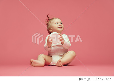 Smiling baby in comfortable onesie sitting on pink background, holding moisturizing skin cream. Cosmetics for babies Smiling baby in comfortable onesie sitting on pink background, holding moisturizing skin cream. Cosmetics for babies 113479587