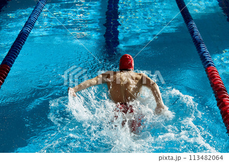 Top view of muscular, athletic young man, swimmer in red cap in motion, showing strength, training, swimming in pool indoors Top view of muscular, athletic young man, swimmer in red cap in motion, showing strength, training, swimming in pool indoors 113482064