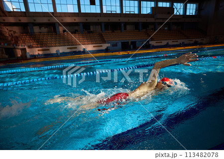 Young man, swimmer in red cap and goggles in motion, training freestyle stroke, swimming in pool indoors 113482078