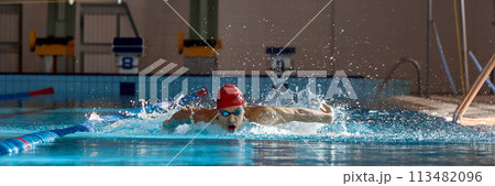 Competitive, muscular young man, swimmer in red cap performs butterfly stroke in pool, training indoors 113482096