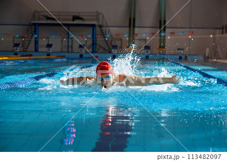 Competitive, muscular young man, swimmer in red cap performs butterfly stroke in pool, training indoors 113482097