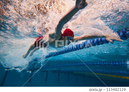 Strength and resilience. Young man, swimmer in red cap and goggles swimming, training in pool indoors. Strength and resilience. Young man, swimmer in red cap and goggles swimming, training in pool indoors. 113482129