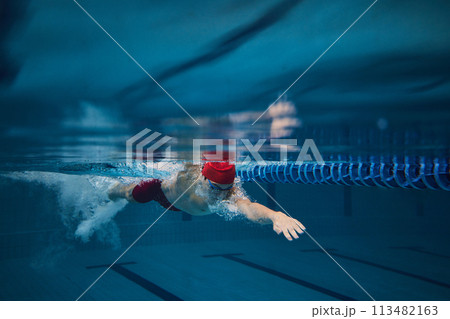 Concentration. Competitive young man, swimming athlete in goggles and red cap in motion, training in pool indoors. Concentration. Competitive young man, swimming athlete in goggles and red cap in motion, training in pool indoors. 113482163