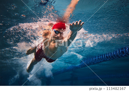 Resilience and strength. Concentrated young man in red cap and goggles in motion, swimming in pool, training. 113482167