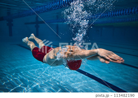 Dynamic image of athletic young guy, swimmer in cap in motion underwater, training in swimming pool. Bubbles, speed. 113482190