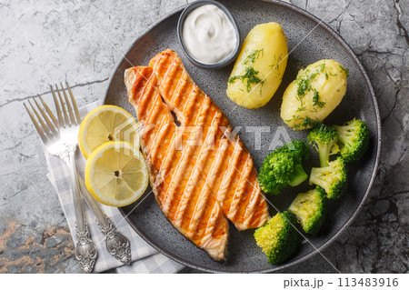 Delicious grilled salmon steak with boiled new potatoes, broccoli, lemon and cream sauce close-up in a plate. Horizontal top view Delicious grilled salmon steak with boiled new potatoes, broccoli, lemon and cream sauce close-up in a plate. Horizontal top view 113483916