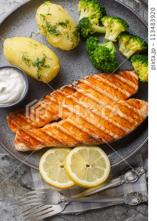 Portion of grilled salmon with boiled potatoes, broccoli, lemon, herbs and cream sauce close-up in a plate on the table. Vertical top view 113483920