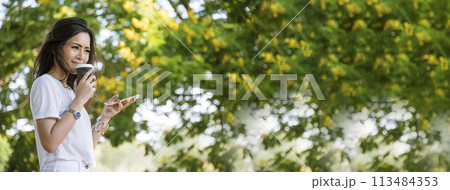 Banner Woman use smartphone drinking coffee hand holding hot disposable cup in green park. Web Banner Happy Relax asian woman smiling face at outdoors garden. Young women enjoy nature with copy space 113484353