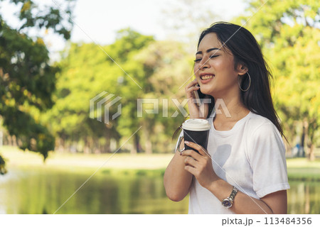 Asian Woman use smartphone drinking coffee hand holding hot disposable cup in green park. Banner Happy Relax asian woman smiling face at outdoors garden. Young women enjoy nature with copy space Asian Woman use smartphone drinking coffee hand holding hot disposable cup in green park. Banner Happy Relax asian woman smiling face at outdoors garden. Young women enjoy nature with copy space 113484356