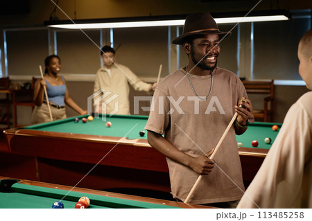 Waist up portrait of African American adult man chatting with friend and smiling while enjoying game of pool together in low light copy space 113485258