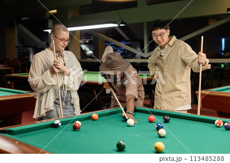 Waist up portrait of diverse group of young people playing billiards together at pool table in underground club watching friend taking shot 113485288