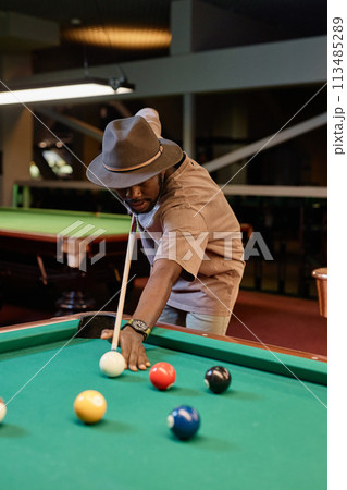Vertical portrait of Black man wearing har and playing pool at billiards table in low lighting 113485289