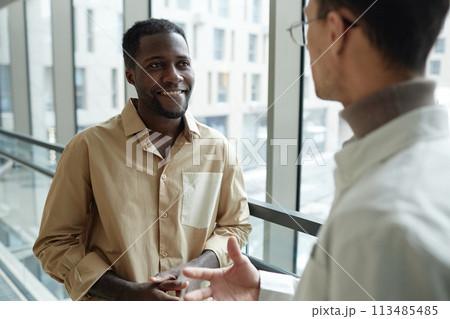 Waist up portrait of smiling Black man talking to colleague standing by window in office 113485485