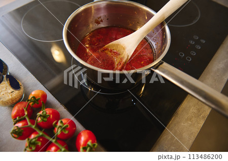 View from above wooden spoon inside a steel pan with boiling homemade sauces of ripe organic juicy red tomatoes. branch of fresh red tomato cherry on the kitchen counter near a black induction cooker View from above wooden spoon inside a steel pan with boiling homemade sauces of ripe organic juicy red tomatoes. branch of fresh red tomato cherry on the kitchen counter near a black induction cooker 113486200
