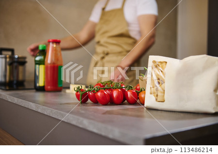 Selective focus on the bunch of red ripe organic tomato cherry and a paper packet with Italian pasta, against blurred male chef standing at induction stove, adjusting program while cooking dinner in 113486214