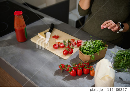 Close-up woman standing at kitchen counter with a fresh bunch of tomato cherry, arugula leaves and Italian pasta in paper packet. Healthy eating, culinary, diet concept. Copy advertising space Close-up woman standing at kitchen counter with a fresh bunch of tomato cherry, arugula leaves and Italian pasta in paper packet. Healthy eating, culinary, diet concept. Copy advertising space 113486237