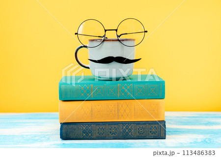 Ceramic cup of coffee with a felt moustache and glasses on the background of a stack of books. Concept of Father's day 113486383