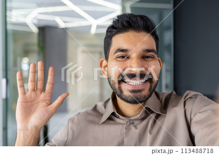 Cheerful businessman engaging in a video call while waving and smiling at the camera, set in a modern office environment. 113488718