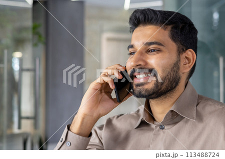 Smiling young man engaged in a phone conversation while in a modern office. This image captures his confidence and pleasant demeanor in a professional environment. 113488724